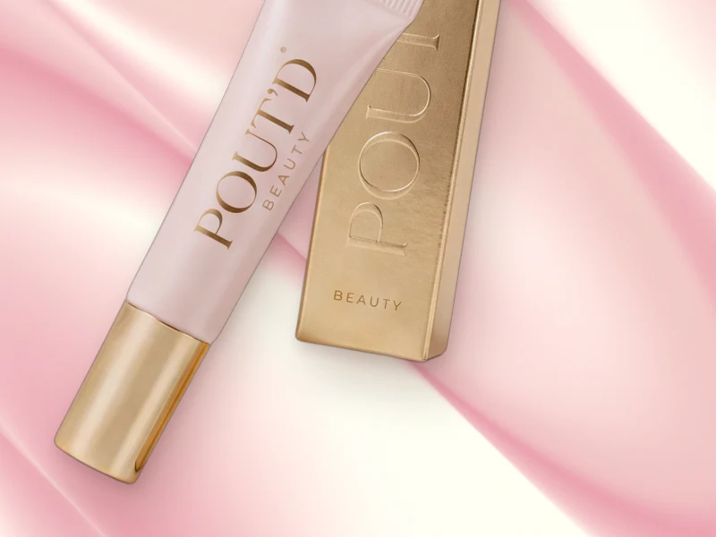 POUT'D lip plumper tube beside its gold box on pink silk, gold cap forward, the $25 buzz-not-burn plumper made for a gentle warmth, not a burn.
